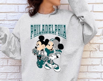 May include: A light grey crewneck sweatshirt featuring a graphic print of two cartoon mice dressed in teal and white sports uniforms. The text PHILADELPHIA is printed in a large, arched teal font above the characters, who are holding pom-poms.