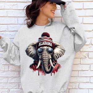 May include: A light gray crewneck sweatshirt featuring a graphic print of an elephant wearing dark sunglasses and a maroon and white knit beanie with the word ALABAMA printed on the front. The elephant is also wearing a maroon hooded sweatshirt.