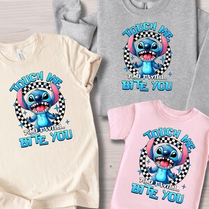 May include: A collection of t-shirts and a sweatshirt featuring a cartoon character. Each shirt displays the text "TOUCH ME AND I WILL BITE YOU" with a blue alien character in a black and white checkered circle. The shirts are in beige, gray, and pink.