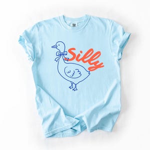 May include: Light blue t-shirt with a cartoon duck illustration and the word "Silly" in red script. The duck is outlined in blue with a bow tie. The shirt is a casual, unisex style.