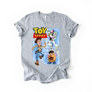 May include: A heather gray short-sleeve t-shirt featuring a graphic print of the Toy Story logo and characters Woody, Buzz Lightyear, and Mr. Potato Head. The design is set against a light blue background with white star accents on the front.
