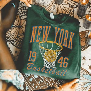 May include: A dark green crewneck sweatshirt featuring the orange text NEW YORK, the number 1946, and the word Basketball. A graphic of a basketball hoop with a net and a basketball inside is centered between the numbers 19 and 46 on the front.