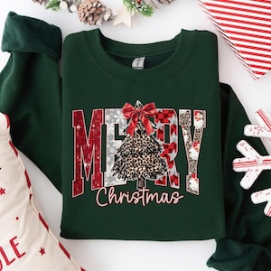 May include: A dark green crewneck sweatshirt featuring the text MERRY Christmas in a stylized font. The letters are filled with red plaid, silver snowflakes, and leopard print patterns. A leopard print Christmas tree with a red bow sits in the center.