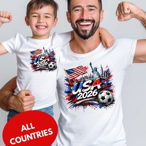 A man and a young boy wearing matching white t-shirts with a graphic design featuring the text "USA 2026" and an American flag, along with a soccer ball.