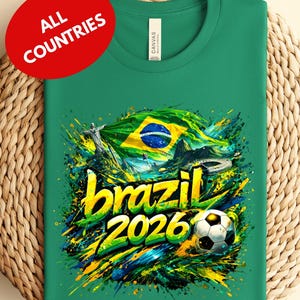 A green t-shirt with a vibrant graphic design featuring the text "brazil 2026" and a soccer ball, surrounded by splashes of color and the Brazilian flag.
