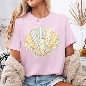 May include: A light pink t-shirt featuring a large, central graphic of a seashell composed of various patchwork patterns. The shell design incorporates yellow and blue stripes, yellow gingham checks, and small yellow floral prints on a white background.