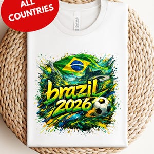 A white t-shirt with a colorful graphic design featuring the text "brazil 2026" and a soccer ball, surrounded by splashes of green, yellow, and blue colors.