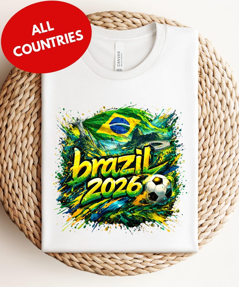A white t-shirt with a colorful graphic design featuring the text "brazil 2026" and a soccer ball, surrounded by splashes of green, yellow, and blue colors.