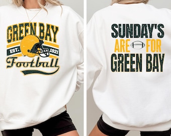 Puede incluir: Una sudadera blanca de cuello redondo con un gráfico de Green Bay Football en la parte delantera, que incluye un casco amarillo y el texto EST. 1921. La parte trasera muestra el texto SUNDAY'S ARE FOR GREEN BAY en verde oscuro y amarillo.