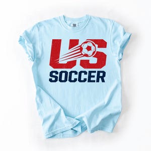 May include: Light blue t-shirt with the text "US SOCCER" in red and navy blue. The design features a soccer ball with speed lines. The shirt is a casual wear item.
