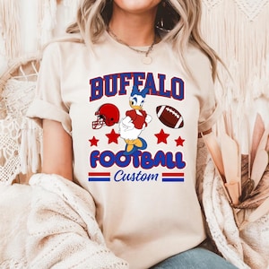 May include: A beige t-shirt featuring a graphic print with the text BUFFALO FOOTBALL Custom in blue and red. The design includes a cartoon duck character, a red football helmet, a brown football, and red stars arranged on the front of the shirt.
