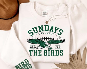 May include: A white long-sleeve sweatshirt featuring a green graphic of an eagle holding a football. The text reads SUNDAYS ARE FOR THE BIRDS in green block letters. The left sleeve features the text BIRD GANG with a small eagle graphic.