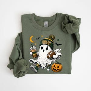 A green sweatshirt with a graphic design depicting a ghost character holding a football and a pumpkin, along with Halloween-themed elements such as bats and a crescent moon.