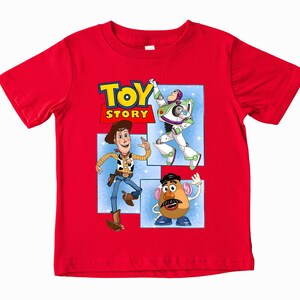May include: A bright red short-sleeved t-shirt featuring a graphic print of the Toy Story logo and characters Woody, Buzz Lightyear, and Mr. Potato Head. The design is set against a light blue background with white star accents on the shirt front.