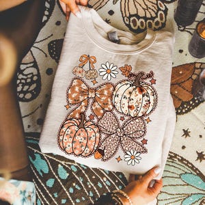 May include: A cream-colored sweatshirt featuring a fall-themed graphic. The design includes pumpkins, bows, daisies, and an acorn, all in shades of orange, brown, and white. The sweatshirt is folded and held by hands, showcasing the detailed print.