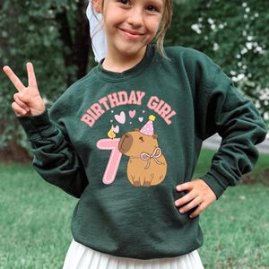 May include: Dark green sweatshirt with the text "BIRTHDAY GIRL" in pink, along with a cartoon capybara wearing a party hat and a pink number 7. The capybara has a pink bow. The sweatshirt is a crew neck.