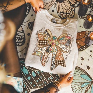 May include: A beige sweatshirt featuring a large bow design composed of various floral and plaid patterns. The text "God is Within Her She Will Not Fall" and "Psalm 46:5" are printed around the bow. The sweatshirt is folded.
