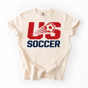 May include: Cream-colored t-shirt with the text "US SOCCER" in red and navy blue. The "US" is stylized with a soccer ball and speed lines. The shirt is a casual, short-sleeved design.