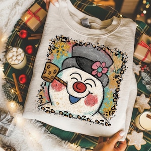 May include: A cream-colored crewneck sweatshirt featuring a graphic of a smiling snowman wearing a gray top hat with a pink flower. The snowman has a red nose, a blue tongue, and holds a brown mallet. The design is framed by a leopard print border.