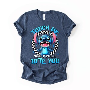 May include: A navy blue t-shirt featuring a cartoon character with blue fur, large ears, and a wide grin. The text "TOUCH ME AND I WILL BITE YOU" is printed in blue, with the character in the center of a black and white checkered circle.