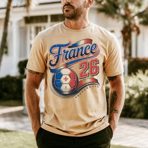K&ouml;nnte beinhalten: Beiges T-Shirt mit dem Wort "France" in blauer und roter Schrift &uuml;ber einem Fu&szlig;ball. Der Fu&szlig;ball ist in den Farben der franz&ouml;sischen Flagge gehalten. Die Nummer "26" ist rot. Eine Reihe von Sternen befindet sich darunter.