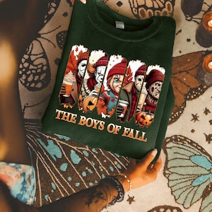 May include: Dark green sweatshirt featuring a graphic of horror movie characters holding fall-themed drinks and pumpkins. The text "THE BOYS OF FALL" is printed in orange. The sweatshirt is a fall-themed item.