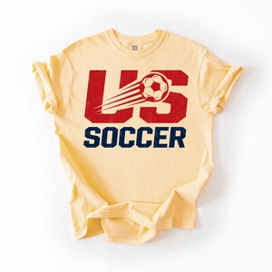 May include: A light yellow t-shirt with the words "US SOCCER" in a distressed red and navy blue font. A soccer ball with motion lines is incorporated into the design. The shirt is made of a soft material.