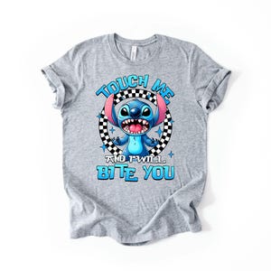 May include: A gray t-shirt featuring a cartoon character with blue fur, large ears, and an open mouth. The text "TOUCH ME AND I WILL BITE YOU" is printed in blue above and below the character.