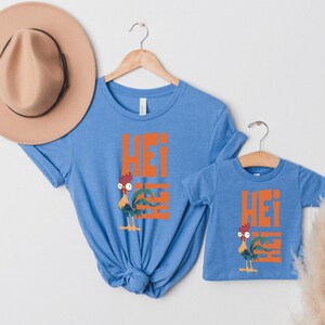 Op de afbeelding: Twee blauwe T-shirts met een cartoon-haan en het woord "Hei" erop gedrukt. Het grotere shirt heeft een haan met een rode kam en rode keellap, het kleinere shirt heeft een haan met een gele kam en gele keellap.