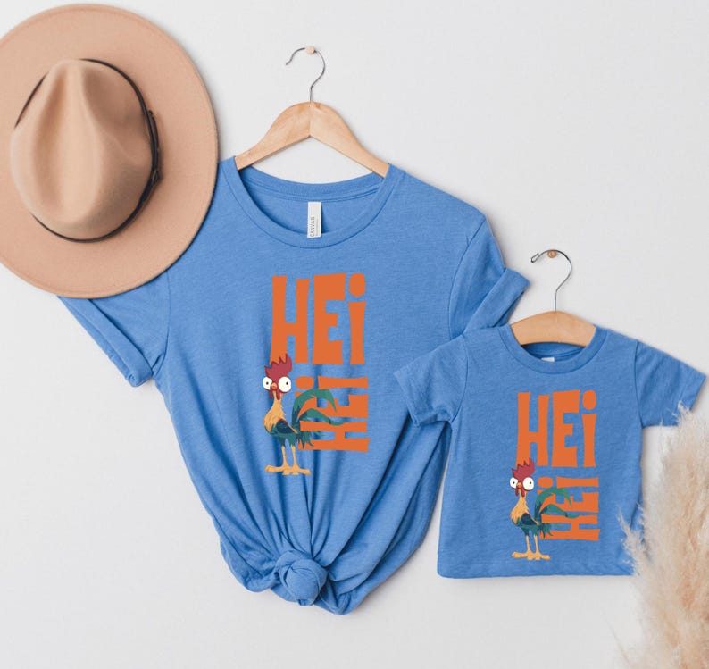Op de afbeelding: Twee blauwe T-shirts met een cartoon-haan en het woord "Hei" erop gedrukt. Het grotere shirt heeft een haan met een rode kam en rode keellap, het kleinere shirt heeft een haan met een gele kam en gele keellap.
