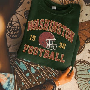 May include: Dark green sweatshirt with the text "WASHINGTON FOOTBALL" in maroon and gold lettering. A maroon football helmet graphic is centered below the word "WASHINGTON". The numbers "19" and "32" are on either side of the helmet.