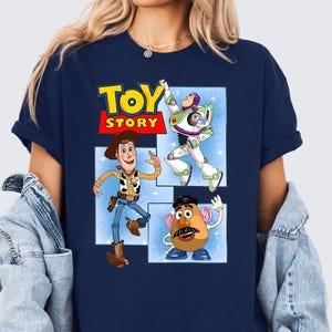 May include: A navy blue short-sleeve t-shirt featuring a graphic print of the Toy Story logo and characters Woody, Buzz Lightyear, and Mr. Potato Head. The characters are displayed in separate rectangular frames against a light blue background.