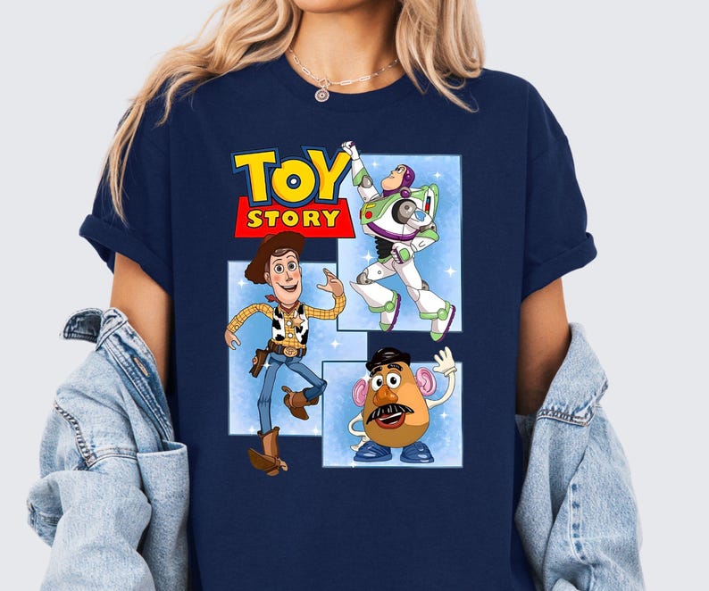 May include: A navy blue short-sleeve t-shirt featuring a graphic print of the Toy Story logo and characters Woody, Buzz Lightyear, and Mr. Potato Head. The characters are displayed in separate rectangular frames against a light blue background.