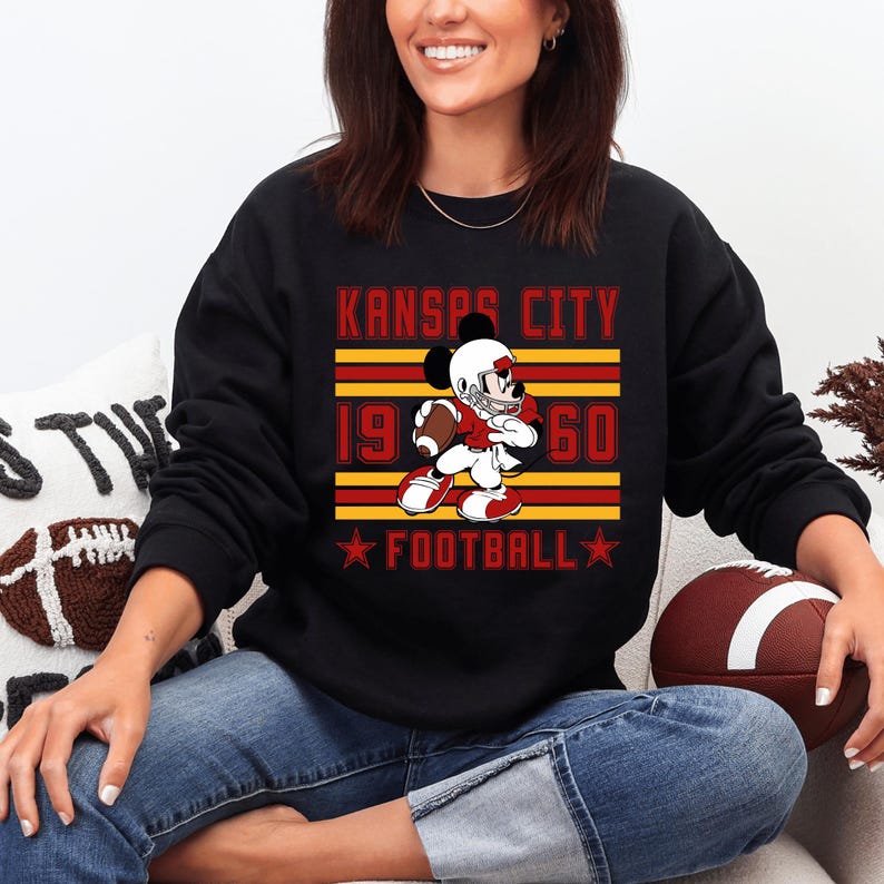 May include: Black sweatshirt featuring a cartoon Mickey Mouse in a football uniform, holding a football. The design includes the text "KANSAS CITY 1960 FOOTBALL" in red and gold. The sweatshirt is worn by a person.