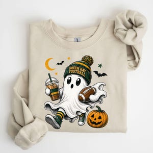 A t-shirt with a Halloween-themed graphic design featuring a ghost character holding a football, a pumpkin, and a cup of a beverage.