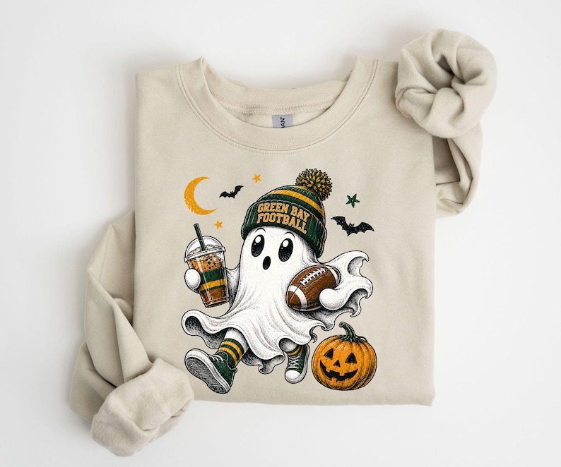 A t-shirt with a Halloween-themed graphic design featuring a ghost character holding a football, a pumpkin, and a cup of a beverage.