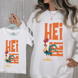Op de afbeelding: Witte T-shirts met een haan-afbeelding en het woord "Hei" in oranje gedrukt. Het grotere shirt heeft een lange mouw en het kleinere shirt heeft een korte mouw.