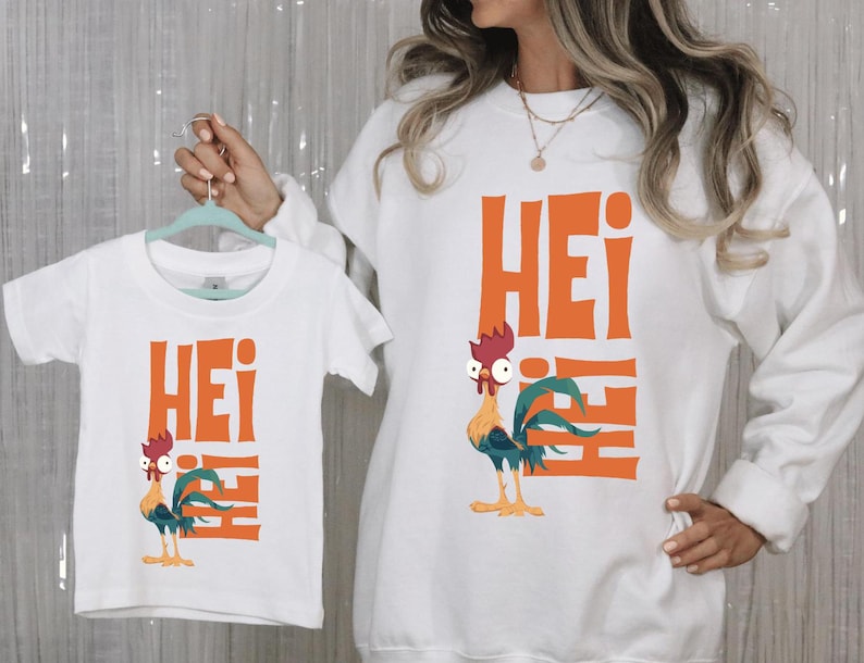Op de afbeelding: Witte T-shirts met een haan-afbeelding en het woord "Hei" in oranje gedrukt. Het grotere shirt heeft een lange mouw en het kleinere shirt heeft een korte mouw.