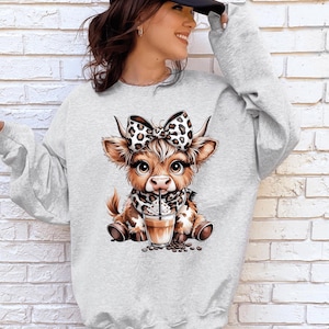 May include: Light gray sweatshirt featuring a cartoon cow with a bow and scarf in a leopard print design. The cow is drinking from a coffee cup with coffee beans around it. The sweatshirt is a casual style.