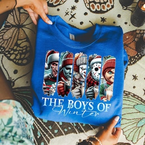 May include: A royal blue sweatshirt featuring a graphic of horror movie characters in winter attire. The text "THE BOYS OF Winter" is printed below the characters. The sweatshirt is held up by a person.