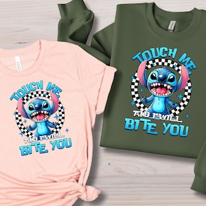 May include: A light pink t-shirt and a green sweatshirt featuring a cartoon character with the text "TOUCH ME AND I WILL BITE YOU". The character is blue with large ears and a wide grin, set within a black and white checkered circle.