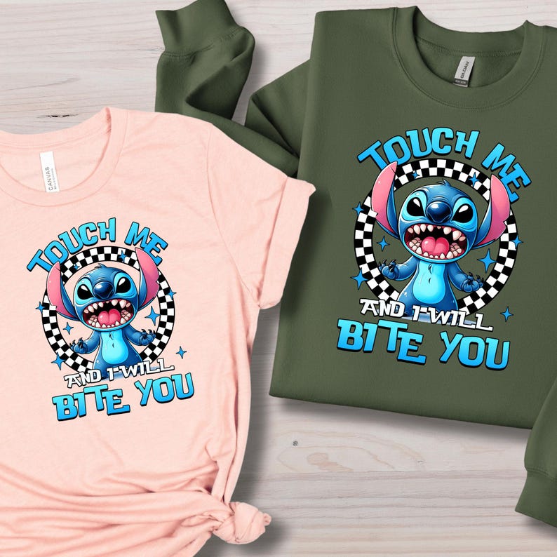 May include: A light pink t-shirt and a green sweatshirt featuring a cartoon character with the text "TOUCH ME AND I WILL BITE YOU". The character is blue with large ears and a wide grin, set within a black and white checkered circle.