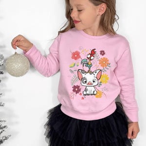 May include: A light pink sweatshirt featuring a graphic of a pig and a rooster with floral accents. The sweatshirt is long-sleeved and worn by a person. The person is wearing a black tulle skirt and holding a silver ornament.