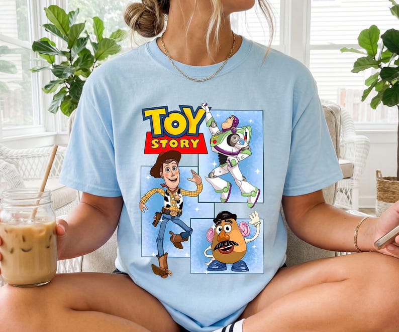 May include: A light blue t-shirt featuring a graphic print of the Toy Story logo in yellow and red, with illustrations of Woody, Buzz Lightyear, and Mr. Potato Head arranged in separate blue rectangular panels on the front of the garment.