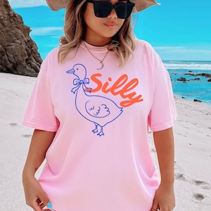 May include: Pink t-shirt with a cartoon duck illustration and the word "Silly" in orange script. The duck is outlined in blue with a blue bow. The shirt is worn by a person wearing sunglasses and a straw hat.