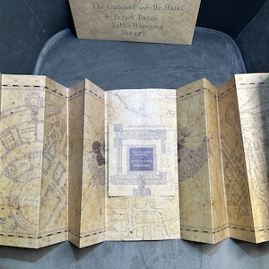 K&ouml;nnte beinhalten: Eine braune und beigefarbene Papierkarte mit einer detaillierten Illustration einer Stadt. Die Karte ist in Abschnitte gefaltet und tr&auml;gt den Text "Marauder's Map" und "Moony Padfoot Wormtail Prongs".