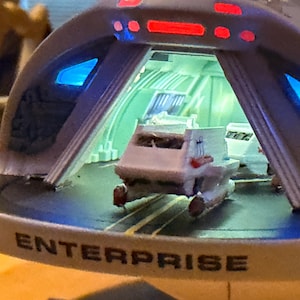 以下が含まれることがあります： ベースに「ENTERPRISE」の文字がプリントされた宇宙船エンタープライズの模型。モデルは白とグレーの外観で、赤と青のライトが付いています。内部には小さなシャトルが見えます。
