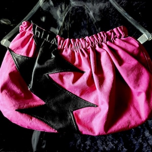 May include: A pink fabric bag with a black lightning bolt design. The bag has a gathered top and is made of a soft, textured material.