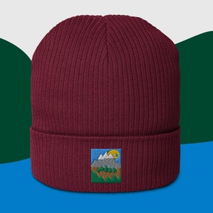 Pode incluir: Um gorro de malha bordô com um pequeno remendo bordado que representa uma cena de montanha com um sol amarelo e árvores verdes.