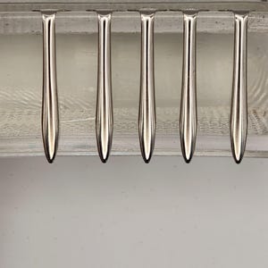 May include: Six shiny, silver-colored forks arranged in a row. The forks have a teardrop-shaped handle and a pointed tip. The forks are set against a clear, textured background.
